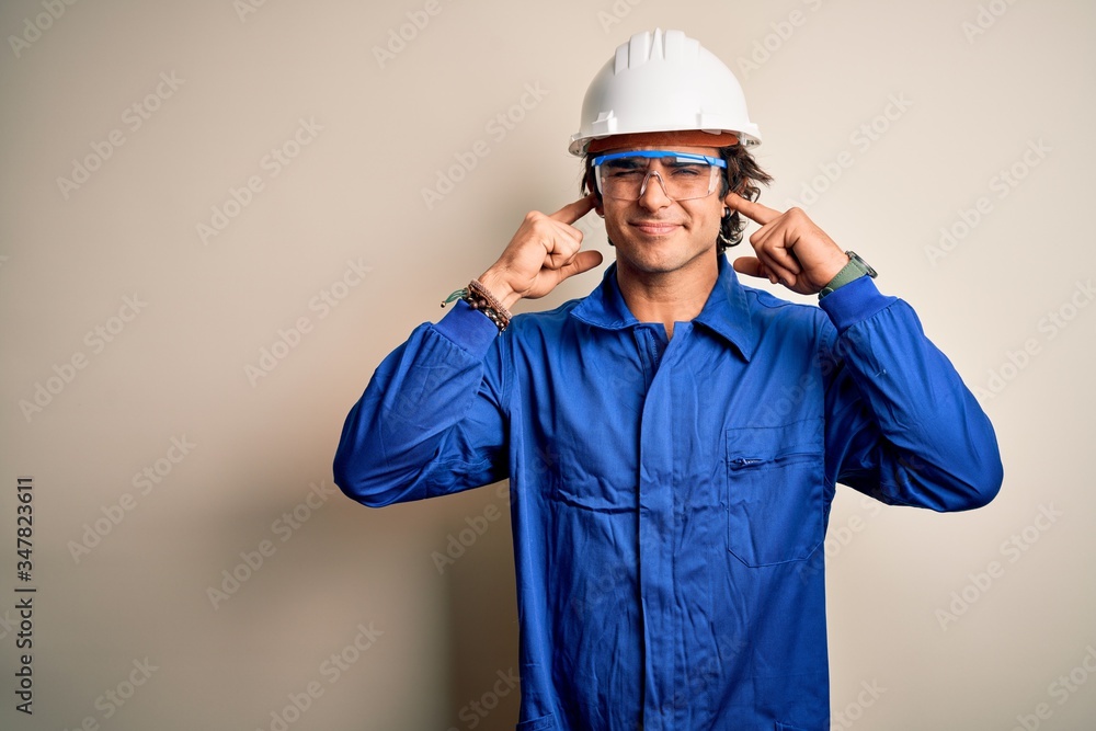 Young constructor man wearing uniform and security helmet over isolated white background covering ears with fingers with annoyed expression for the noise of loud music. Deaf concept.