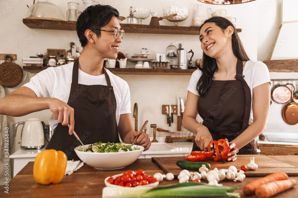 Image of young multicultural couple in aprons laughing and making lunch