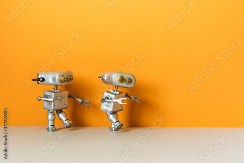 Two metal silver robots are walking. Simplified symbolic toy robotic characters on an orange background. Empty space for text.