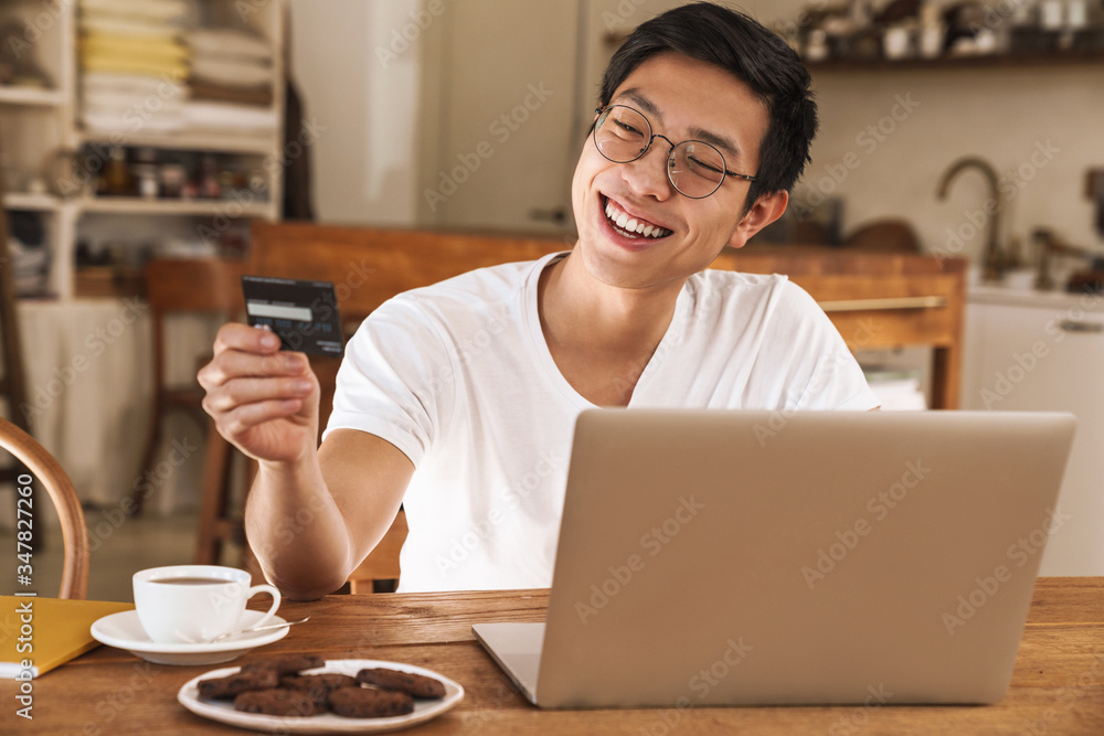 Image of asian man winking while holding credit card and using laptop Image of asian man winking while holding credit card and using laptop