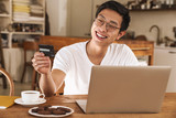 Image of asian man winking while holding credit card and using laptop