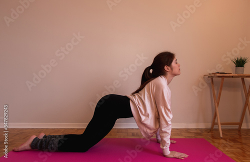 A happy redhead woman wearing pink sweater black leggings and gray legs warmers stretching her body on a pink mat doing a yoga pose with the closed eyes, a plant on the table and a wall background 