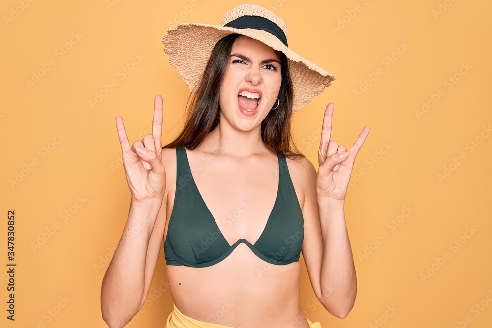 Young beautiful girl wearing swimwear bikini and summer sun hat over yellow background shouting with crazy expression doing rock symbol with hands up. Music star. Heavy concept. Young beautiful girl wearing swimwear bikini and summer sun hat over yellow background shouting with crazy expression doing rock symbol with hands up. Music star. Heavy concept.