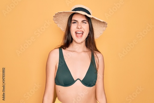 Young beautiful girl wearing swimwear bikini and summer sun hat over yellow background winking looking at the camera with sexy expression  cheerful and happy face.