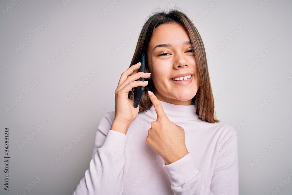 Beautiful brunette girl having conversation talking on the smartphone over white background very happy pointing with hand and finger Beautiful brunette girl having conversation talking on the smartphone over white background very happy pointing with hand and finger