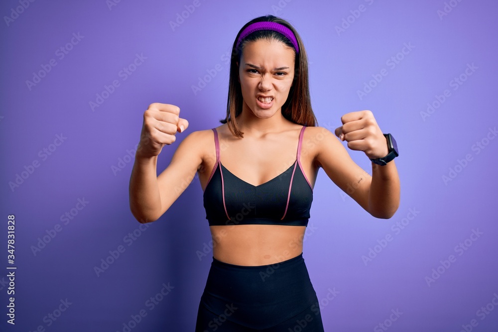Young beautiful sporty girl doing sport wearing sportswear over isolated purple background angry and mad raising fists frustrated and furious while shouting with anger. Rage and aggressive concept. Young beautiful sporty girl doing sport wearing sportswear over isolated purple background angry and mad raising fists frustrated and furious while shouting with anger. Rage and aggressive concept.