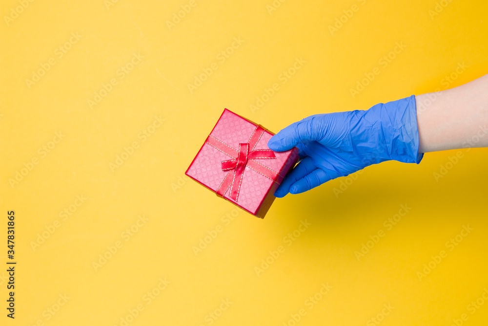 a hand in a blue disposable glove holds a red gift box with a bow from a red ribbon with gold thread, yellow background, copy space
