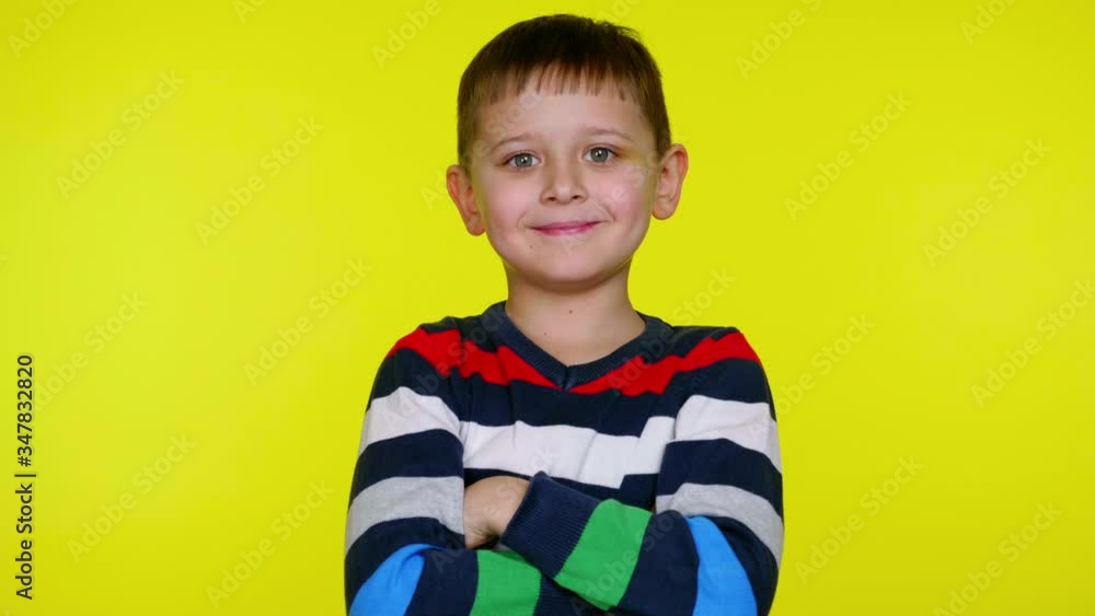 Cheerful little child boy in a multi-colored sweater turns to the camera, crosses arms over chest and smiles on a yellow background with copy space. Place for text or product. 4K slowmotion footage Cheerful little child boy in a multi-colored sweater turns to the camera, crosses arms over chest and smiles on a yellow background with copy space. Place for text or product. 4K slowmotion footage