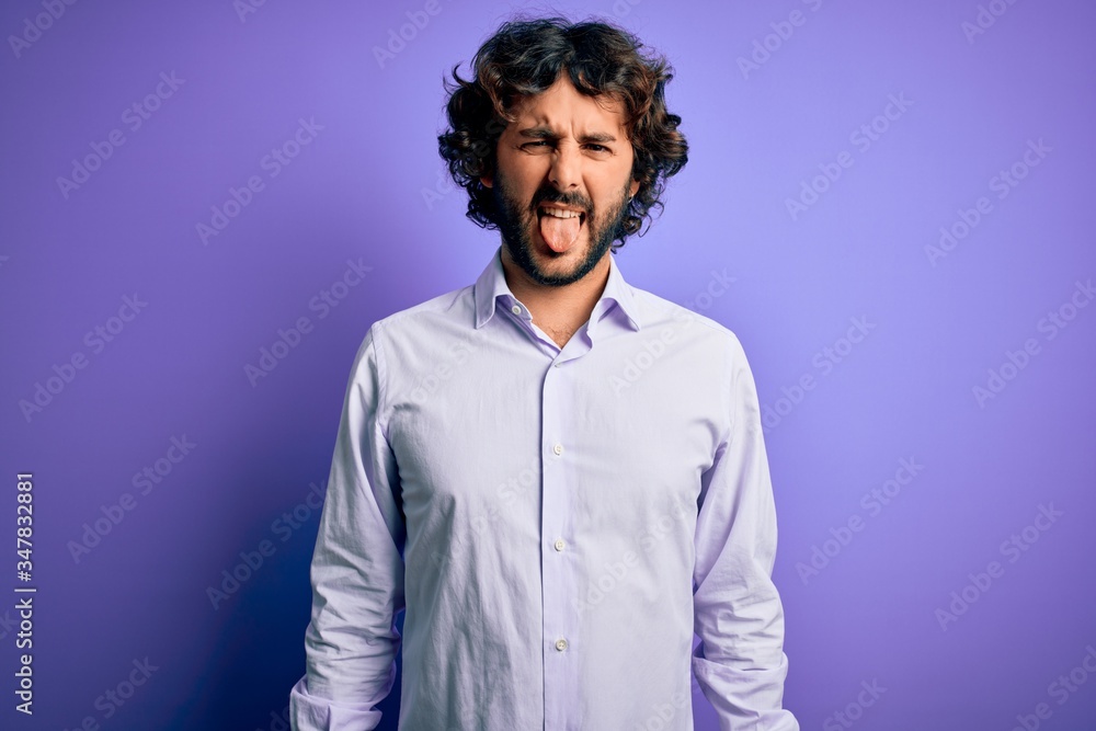 Obraz premium Young handsome business man with beard wearing shirt standing over purple background sticking tongue out happy with funny expression. Emotion concept.