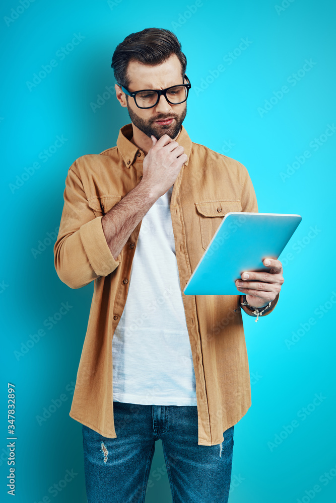 Thoughtful young man in smart casual wear using digital tablet and keeping hand on chin while standing against blue background Thoughtful young man in smart casual wear using digital tablet and keeping hand on chin while standing against blue background