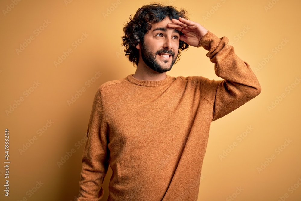 Young handsome man with beard wearing casual sweater standing over yellow background very happy and smiling looking far away with hand over head. Searching concept.