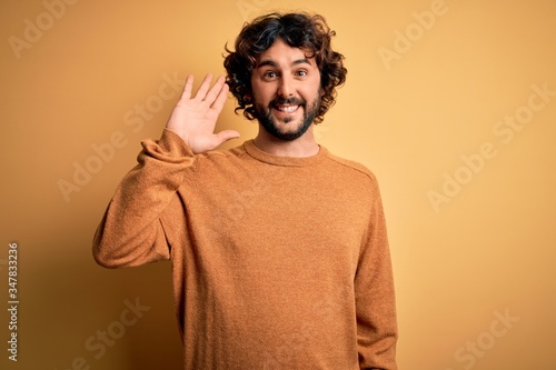 Young handsome man with beard wearing casual sweater standing over yellow background Waiving saying hello happy and smiling, friendly welcome gesture