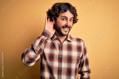 Young handsome man with beard wearing casual shirt standing over yellow background smiling with hand over ear listening an hearing to rumor or gossip. Deafness concept.