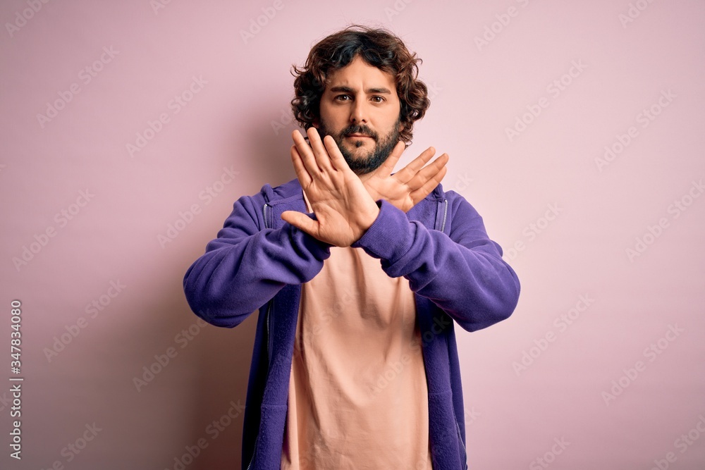 Young handsome sporty man with beard wearing casual sweatshirt over pink background Rejection expression crossing arms and palms doing negative sign, angry face Young handsome sporty man with beard wearing casual sweatshirt over pink background Rejection expression crossing arms and palms doing negative sign, angry face