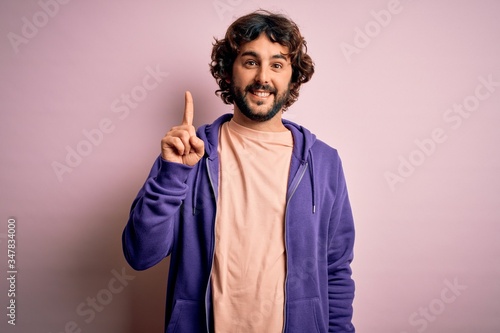 Young handsome sporty man with beard wearing casual sweatshirt over pink background pointing finger up with successful idea. Exited and happy. Number one.