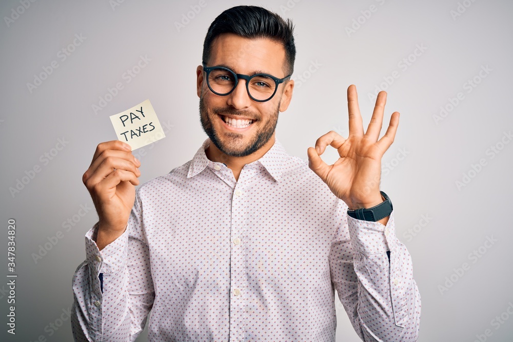 Young handsome man holding reminder paper with pay taxes message over white background doing ok sign with fingers, excellent symbol Young handsome man holding reminder paper with pay taxes message over white background doing ok sign with fingers, excellent symbol