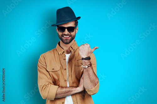 Charming young man smiling and pointing copy space while standing against blue background