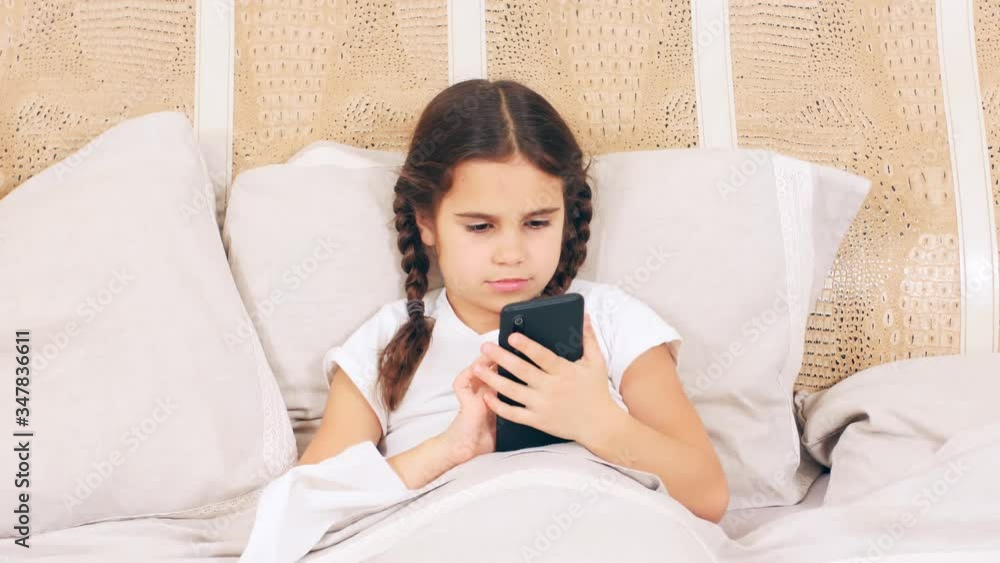 Cute baby girl lies at home in bed, is sick and plays on the phone Cute baby girl lies at home in bed, is sick and plays on the phone