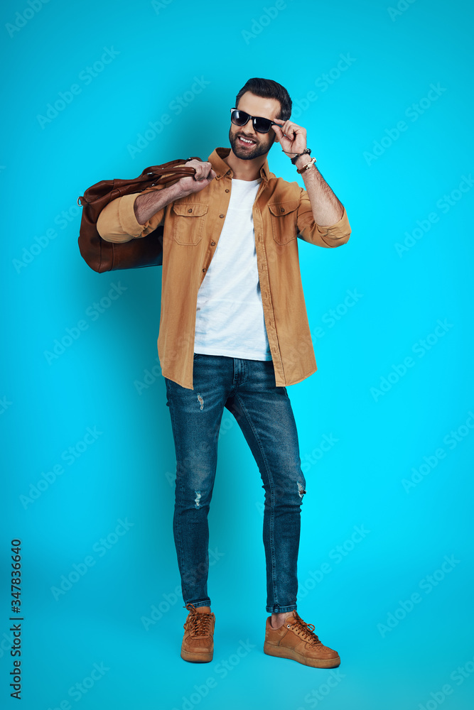 Full length of handsome young man smiling and adjusting eyewear while standing against blue background Full length of handsome young man smiling and adjusting eyewear while standing against blue background