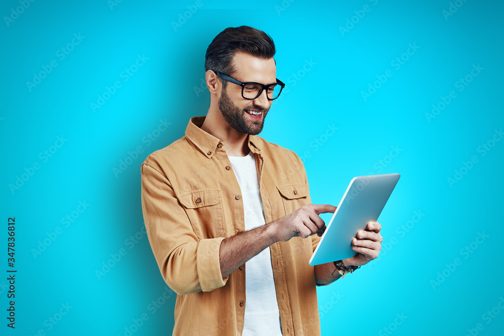 Busy young man in smart casual wear using digital tablet while standing against blue background