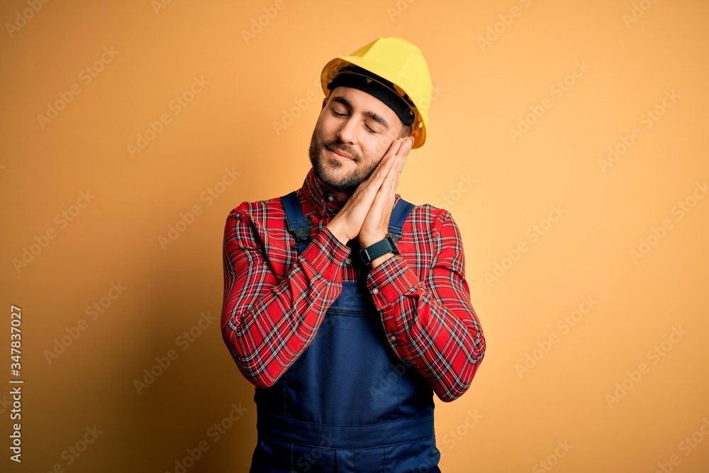 Young builder man wearing construction uniform and safety helmet over yellow isolated background sleeping tired dreaming and posing with hands together while smiling with closed eyes.