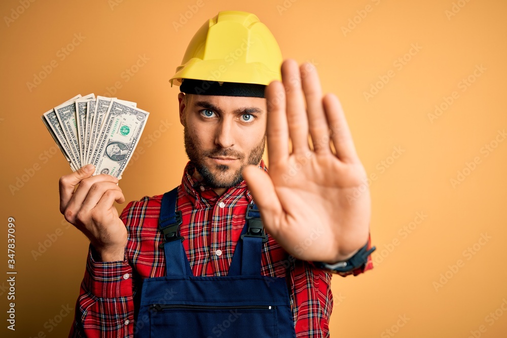 Young builder man wearing safety helmet holding dollars as payment over yellow background with open hand doing stop sign with serious and confident expression, defense gesture Young builder man wearing safety helmet holding dollars as payment over yellow background with open hand doing stop sign with serious and confident expression, defense gesture