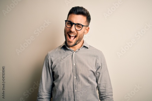 Young handsome man wearing elegant shirt and glasses over isolated white background winking looking at the camera with sexy expression, cheerful and happy face. © Krakenimages.com