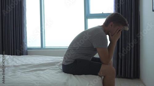 Depressed young man sitting on edge of bed at home or in hotel room.