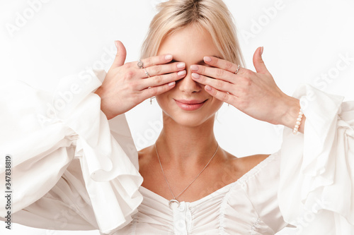 Image of joyful blonde woman in earrings smiling and covering her eyes