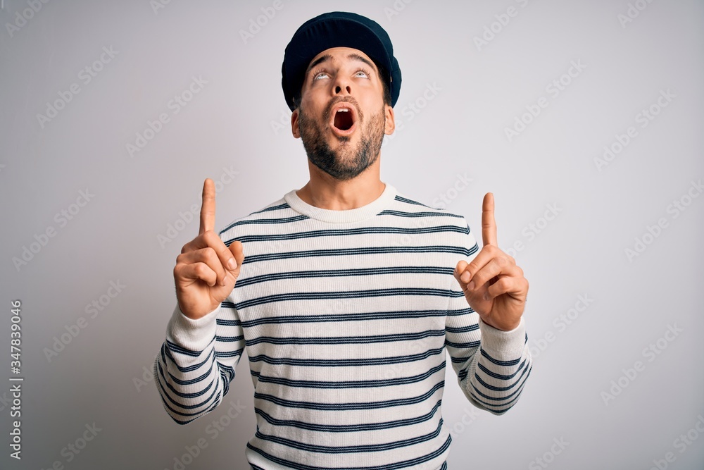 Young handsome sailor man with beard wearing navy striped uniform and captain hat amazed and surprised looking up and pointing with fingers and raised arms. Young handsome sailor man with beard wearing navy striped uniform and captain hat amazed and surprised looking up and pointing with fingers and raised arms.