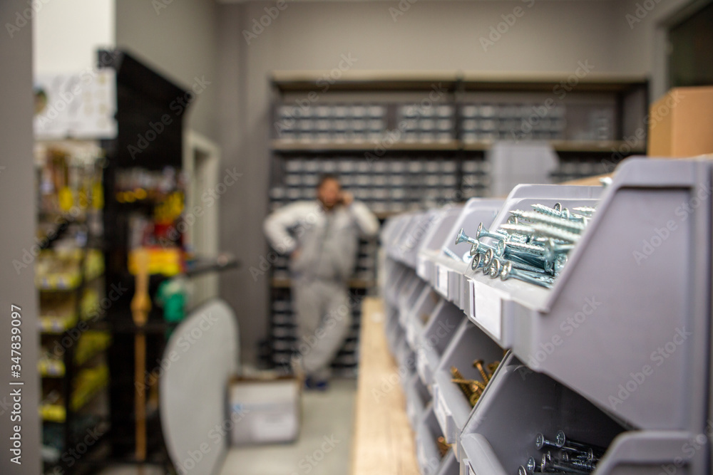 Focus on white metal screws in a plastic box. Behind is a man talking on the phone. Screws in a hardware store and building supplies.