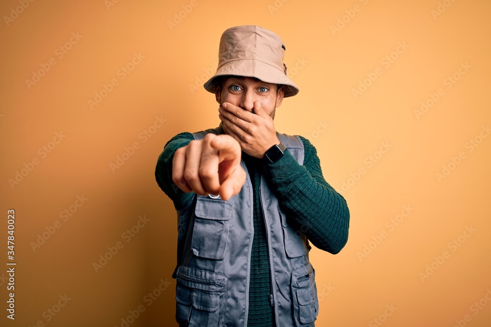 Handsome tourist man with beard on vacation wearing explorer hat over yellow background laughing at you, pointing finger to the camera with hand over mouth, shame expression