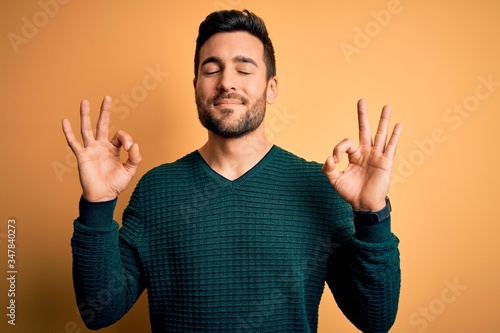 Young handsome man with beard wearing casual sweater standing over yellow background relax and smiling with eyes closed doing meditation gesture with fingers. Yoga concept. © Krakenimages.com