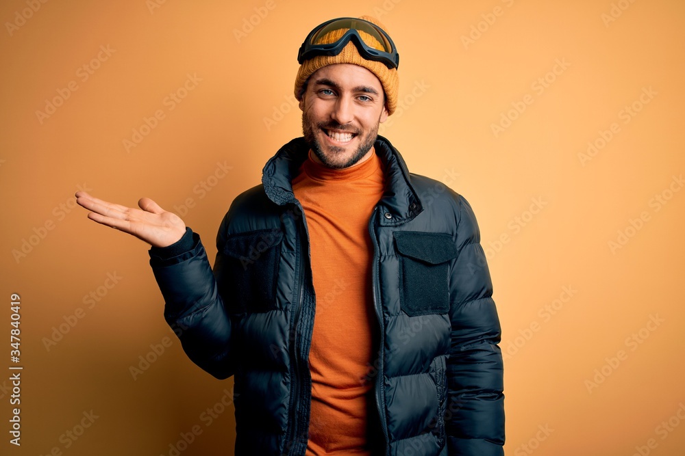 Young handsome skier man with beard wearing snow sportswear and ski goggles smiling cheerful presenting and pointing with palm of hand looking at the camera.