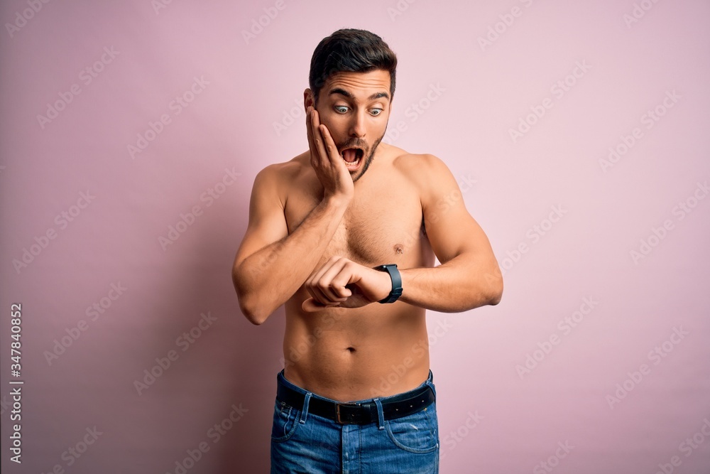 Young handsome strong man with beard shirtless standing over isolated pink background Looking at the watch time worried, afraid of getting late
