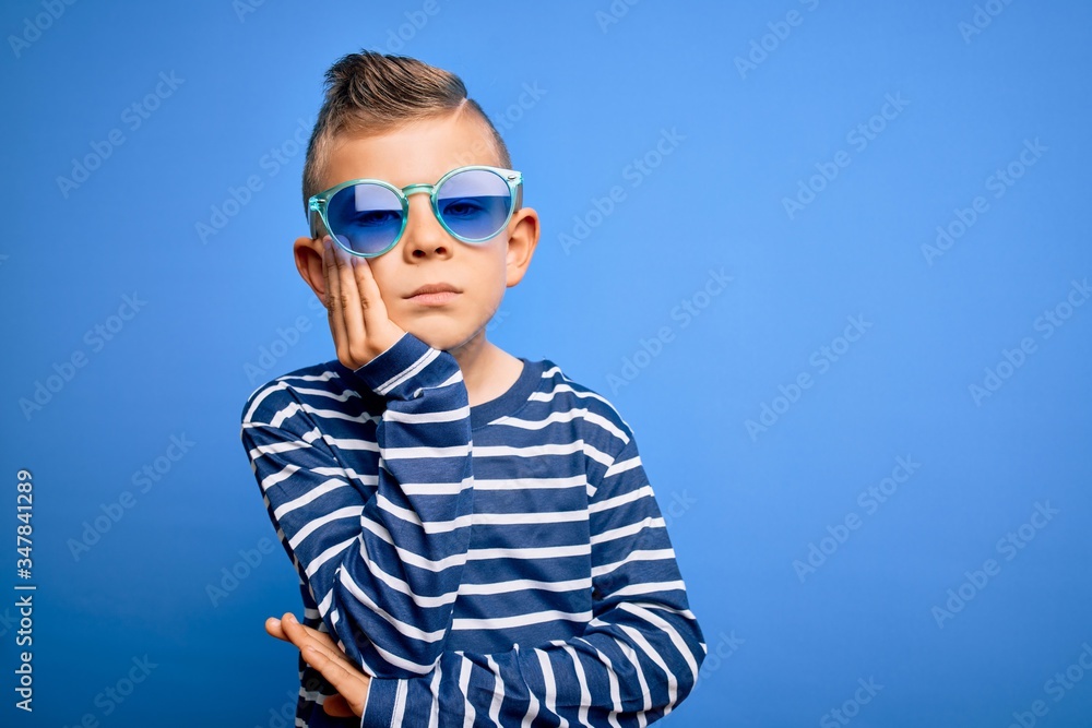 Young little caucasian kid with blue eyes standing wearing sunglasses over blue background thinking looking tired and bored with depression problems with crossed arms.