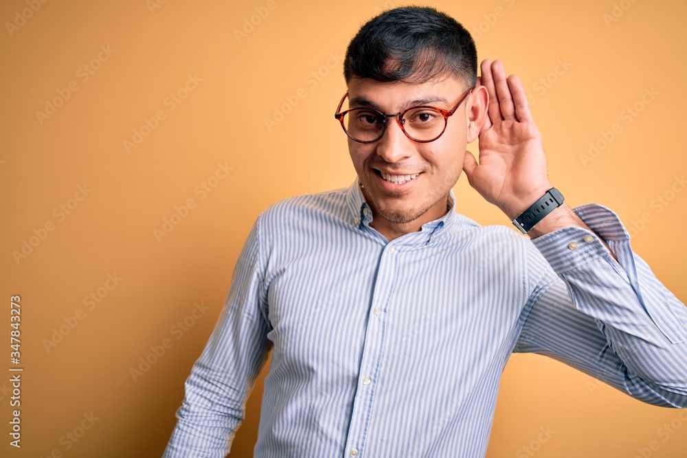Young handsome hispanic business man wearing nerd glasses over yellow background smiling with hand over ear listening an hearing to rumor or gossip. Deafness concept. Young handsome hispanic business man wearing nerd glasses over yellow background smiling with hand over ear listening an hearing to rumor or gossip. Deafness concept.