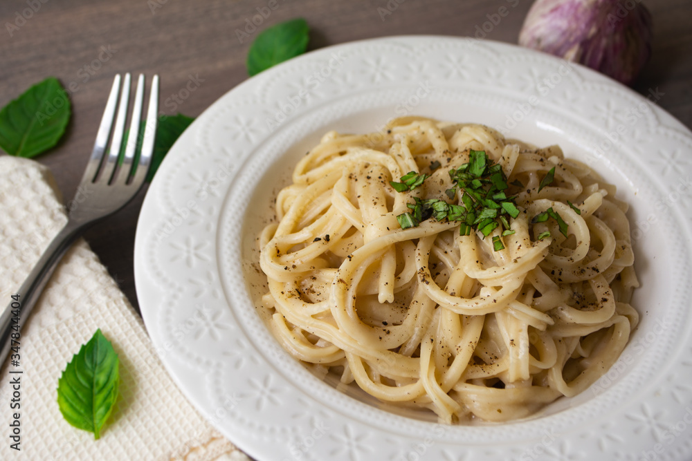 A vegan version of traditional Italian pasta fettuccine alfredo with creamy white sauce garnished with basil on a wooden surface with a fork, napkin, garlic and basil leaves