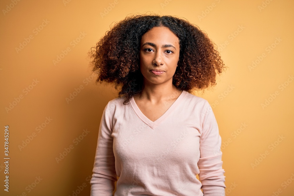 Young african american woman with afro hair wearing casual sweater over yellow background with serious expression on face. Simple and natural looking at the camera. Young african american woman with afro hair wearing casual sweater over yellow background with serious expression on face. Simple and natural looking at the camera.