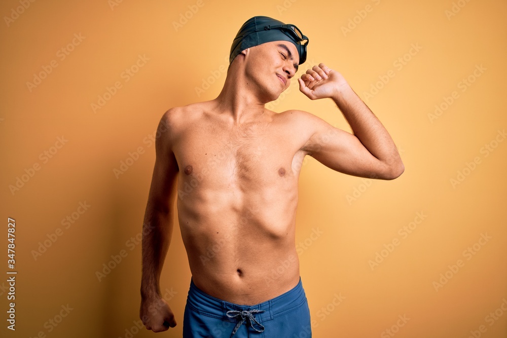 Young handsome man shirtless wearing swimsuit and swim cap over isolated yellow background stretching back, tired and relaxed, sleepy and yawning for early morning