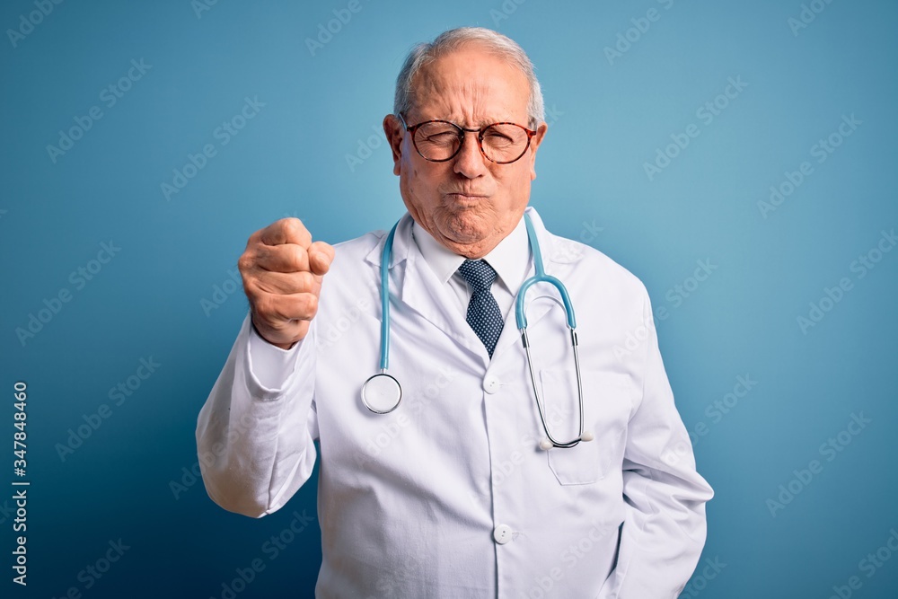 Senior grey haired doctor man wearing stethoscope and medical coat over blue background angry and mad raising fist frustrated and furious while shouting with anger. Rage and aggressive concept. Senior grey haired doctor man wearing stethoscope and medical coat over blue background angry and mad raising fist frustrated and furious while shouting with anger. Rage and aggressive concept.