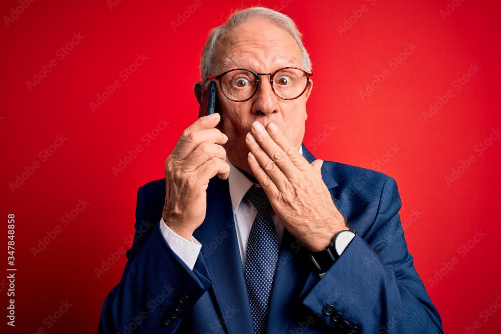 Grey haired senior business man having a conversation talking on smartphone over red background cover mouth with hand shocked with shame for mistake, expression of fear, scared in silence, secret Grey haired senior business man having a conversation talking on smartphone over red background cover mouth with hand shocked with shame for mistake, expression of fear, scared in silence, secret
