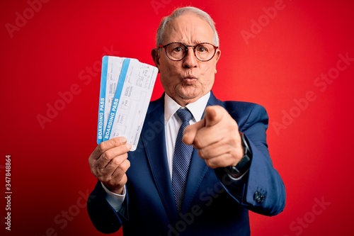 Senior grey haired business man holding airplane boarding pass over red background pointing with finger to the camera and to you, hand sign, positive and confident gesture from the front