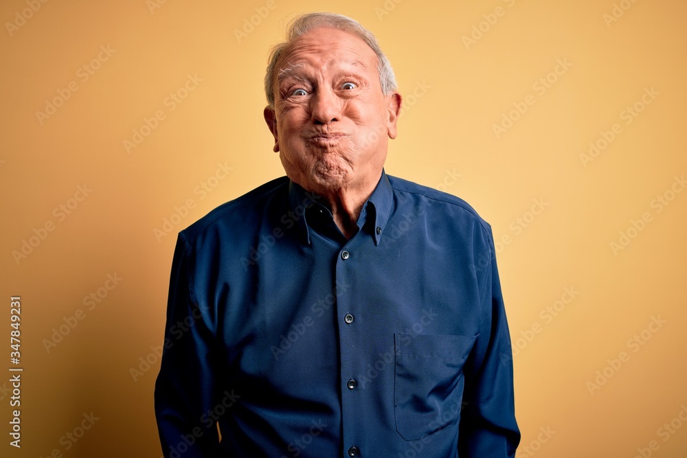 Grey haired senior man wearing casual blue shirt standing over yellow background puffing cheeks with funny face. Mouth inflated with air, crazy expression.