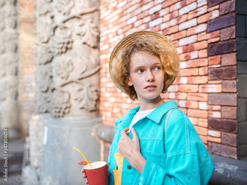 young cheerful curly redhead woman in blue jeans jacket and straw hat (traveler) holding cup of coffee and surprised by landmarks. Fun, summer, travel, fashion, youth concept