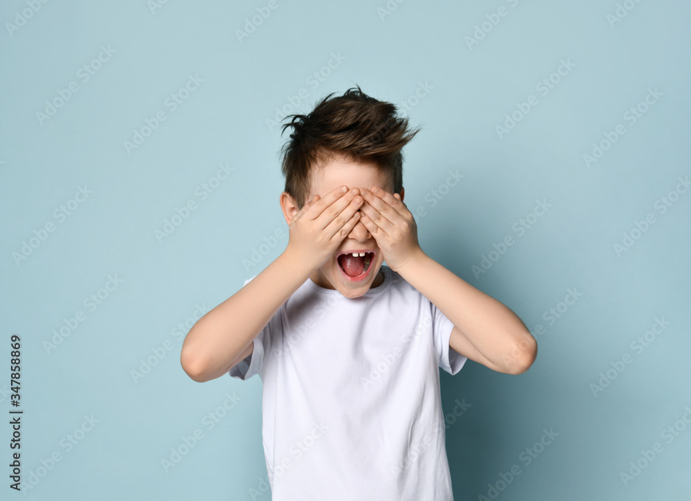 Cute schoolboy standing with hands over eyes playing hide and seek, counting loudly before going after friends. Portrait isolated on light blue Cute schoolboy standing with hands over eyes playing hide and seek, counting loudly before going after friends. Portrait isolated on light blue