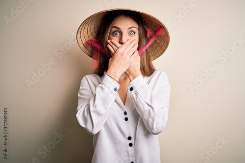 Young beautiful redhead woman wearing asian traditional conical hat over white background shocked covering mouth with hands for mistake. Secret concept.