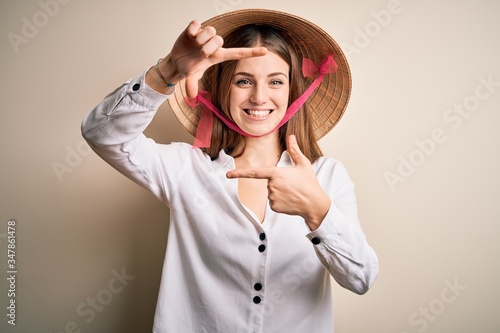 Young beautiful redhead woman wearing asian traditional conical hat over white background smiling making frame with hands and fingers with happy face. Creativity and photography concept.