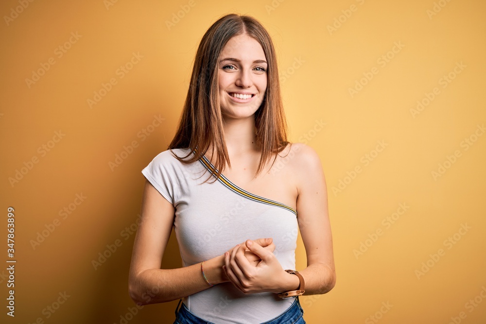 Obraz premium Young beautiful redhead woman wearing casual t-shirt over isolated yellow background with hands together and crossed fingers smiling relaxed and cheerful. Success and optimistic