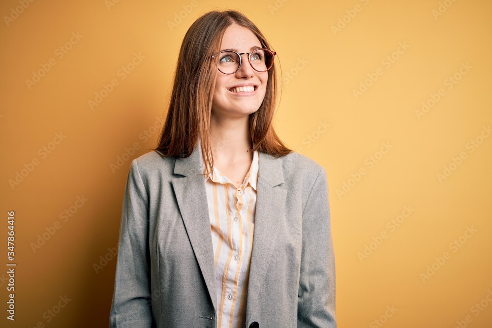 Young beautiful redhead woman wearing jacket and glasses over isolated yellow background looking away to side with smile on face, natural expression. Laughing confident.